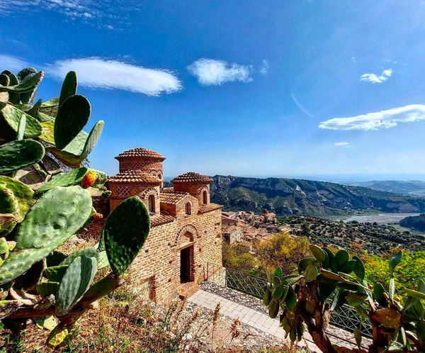 TThe Cattolica di Stilo, a small Byzantine church with distinctive domes, stands amidst lush prickly pear cacti. The terracotta brick structure contrasts with the vast landscape of rolling hills, olive groves, and a winding river in the background. The clear blue sky and scattered clouds add depth to this picturesque Calabrian setting.