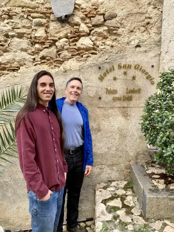 Two men stand in front of a rustic stone wall with a plaque reading "Hotel San Giorgio" and an inscription below. The man on the left, with long hair and wearing a burgundy shirt, smiles warmly, while the older man on the right, dressed in a blue jacket, also smiles. The textured wall and surrounding greenery add to the historical ambiance.