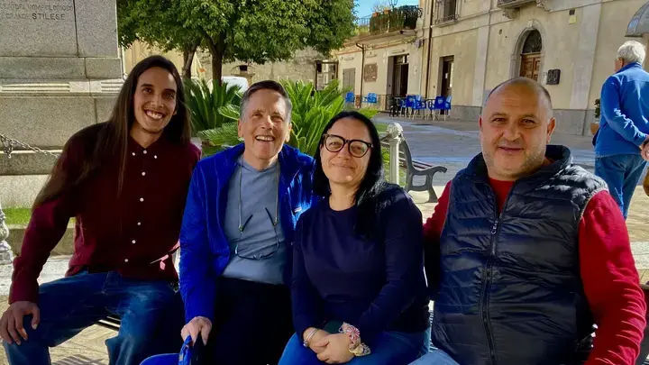 Four people sit together on a bench in a sunlit town square, smiling at the camera. The man on the left, with long hair and a burgundy shirt, sits casually, while the older man next to him wears a blue jacket. A woman in glasses and a dark sweater sits beside another man in a red shirt and black vest. The background features a historic monument, greenery, and a European-style plaza with buildings and outdoor seating.