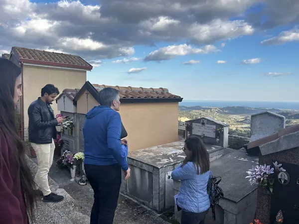 A small group visits a hilltop cemetery in Stilo, surrounded by tombstones and family mausoleums. The location offers a panoramic view of the Calabrian countryside stretching towards the sea, under a sky filled with dramatic clouds. The visitors appear engaged in quiet reflection, adding to the solemn and emotional atmosphere of the moment.