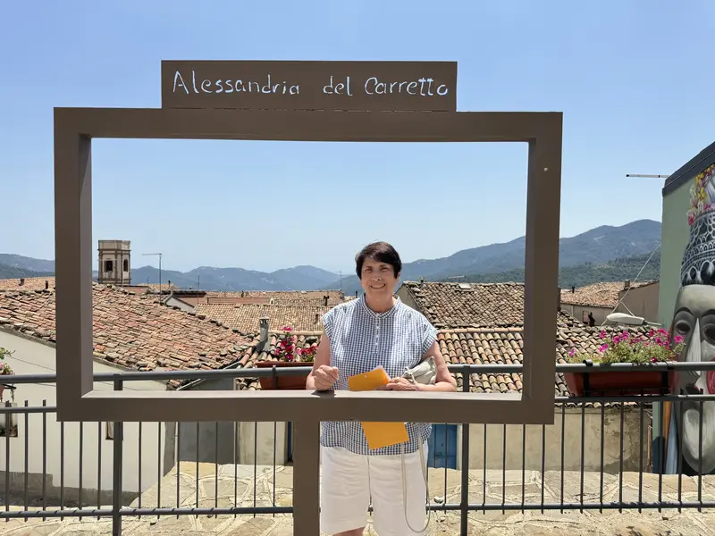 A woman smiles while standing inside a panoramic frame in Alessandria del Carretto, with terracotta rooftops and mountains in the background.